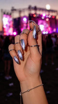 May include: A hand with long, silver chrome nails and multiple silver rings. A delicate silver bracelet adorns the wrist. The background is blurred, suggesting a concert or outdoor event with colorful lights.