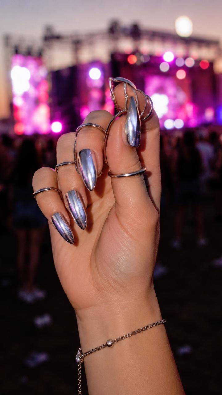 May include: A hand with long, silver chrome nails and multiple silver rings. A delicate silver bracelet adorns the wrist. The background is blurred, suggesting a concert or outdoor event with colorful lights.